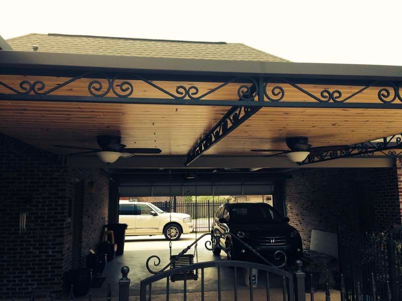Carport with ornate metal trim, parked black vehicle, and a cream car visible beyond the driveway.