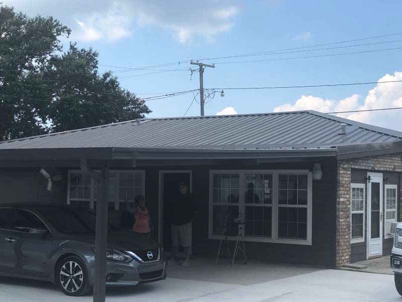 Single-story house with a dark roof, carport, parked car, and cloudy sky.