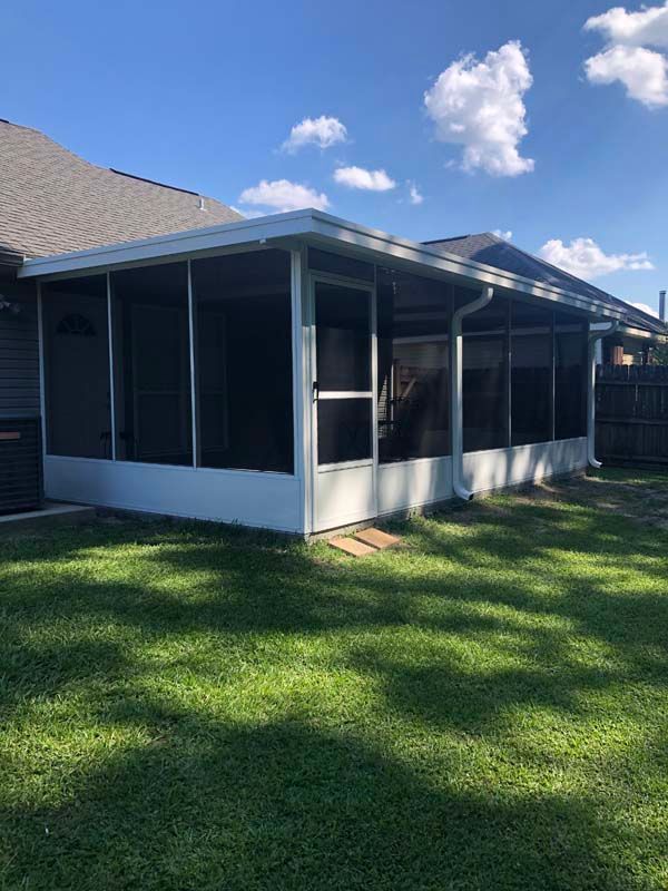 Screened porch with white trim attached to a house, beside a grassy yard under a blue sky.
