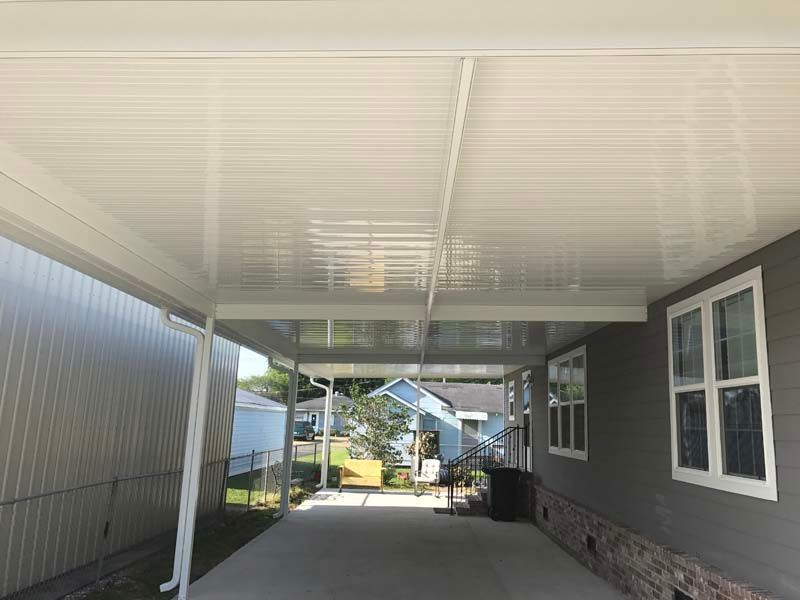 Covered driveway between two houses, with white ceiling and gray siding, leading to a bright yard.