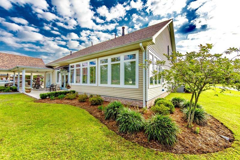 Single-story house with large windows, landscaped garden, and green lawn under a cloudy blue sky