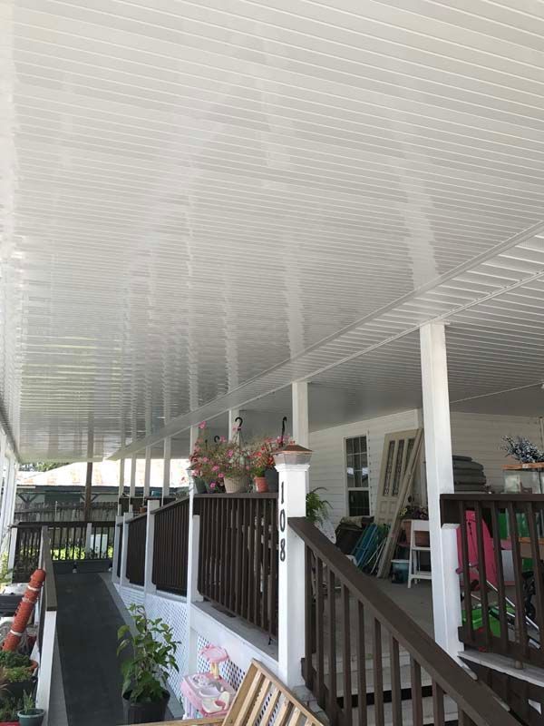 Covered porch with white ceiling, dark railing, hanging plants, and stacked chairs along the side of a house