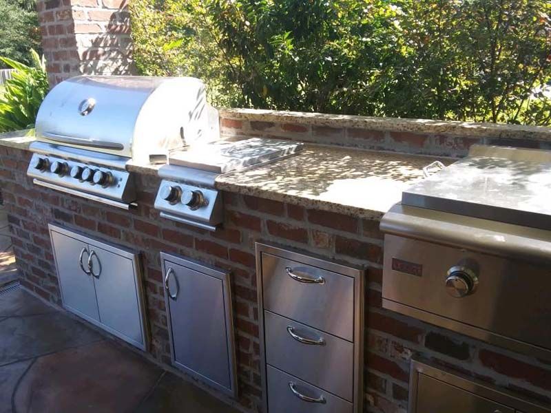 Outdoor brick patio with built-in stainless steel grill, storage doors, and countertop beside greenery