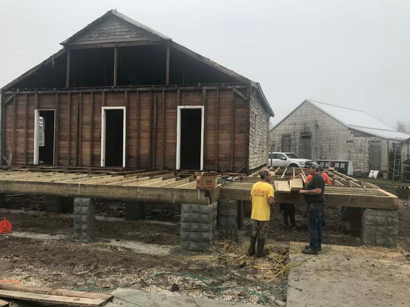 Two workers stand by a raised wooden house under construction on a muddy site.