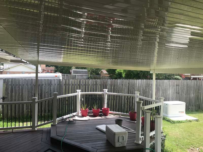 Covered backyard deck with black railing, potted plants, and outdoor furniture beside a fenced lawn.
