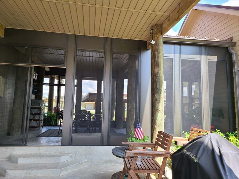 Covered patio with screened doors, wooden chairs, and a grill beside steps leading to the house
