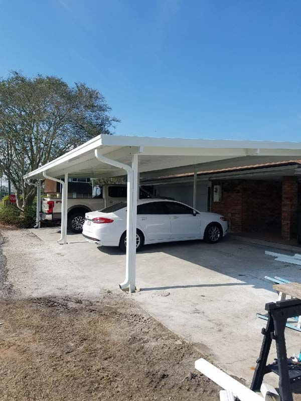 White sedan parked under a carport beside a brick house on a sunny day