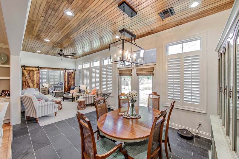 Bright sunroom dining area with round table, white shutters, wood ceiling, and adjacent sitting room