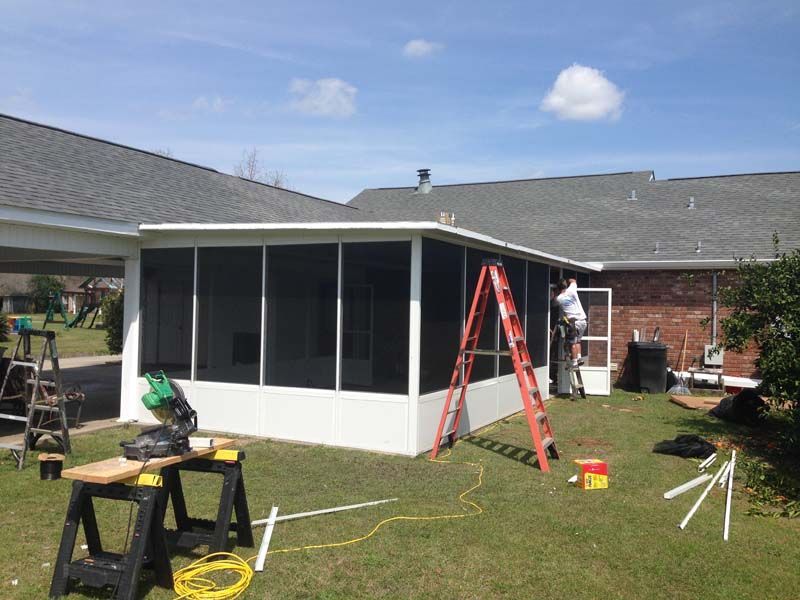 Workers installing screened porch on a house, with ladders, tools, and white framing.