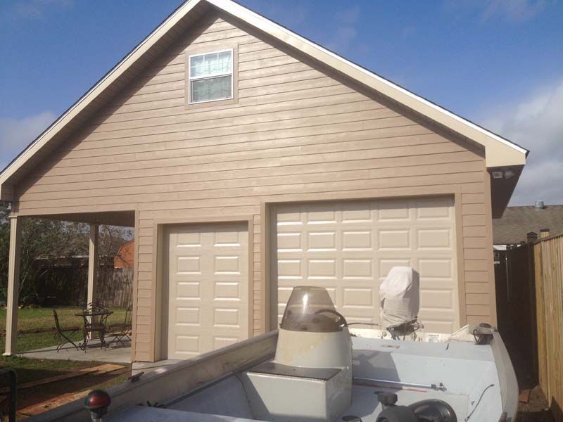 Tan garage with a dormer window, two white doors, and a truck or trailer in front under a blue sky