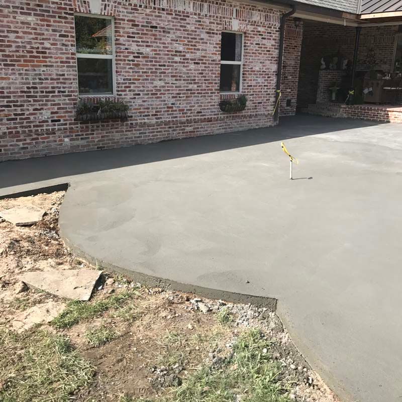 Freshly poured concrete patio beside a brick house, with a small yellow survey marker in the slab