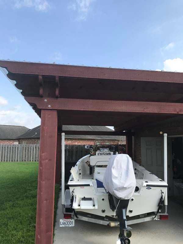 White boat parked under a brown carport in a suburban yard, covered at the stern.
