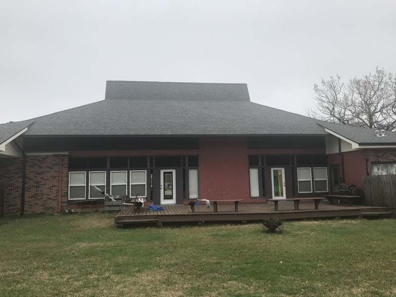 Single-story brick building with dark roof, front windows, and a wooden deck on a grassy lawn under cloudy sky