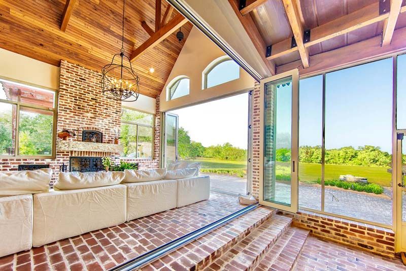 Bright open living room with exposed wood beams, brick fireplace, large windows, and view of a green landscape