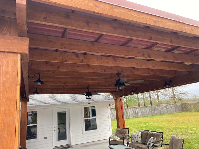Wooden covered patio with ceiling fans overlooking a white house and green yard