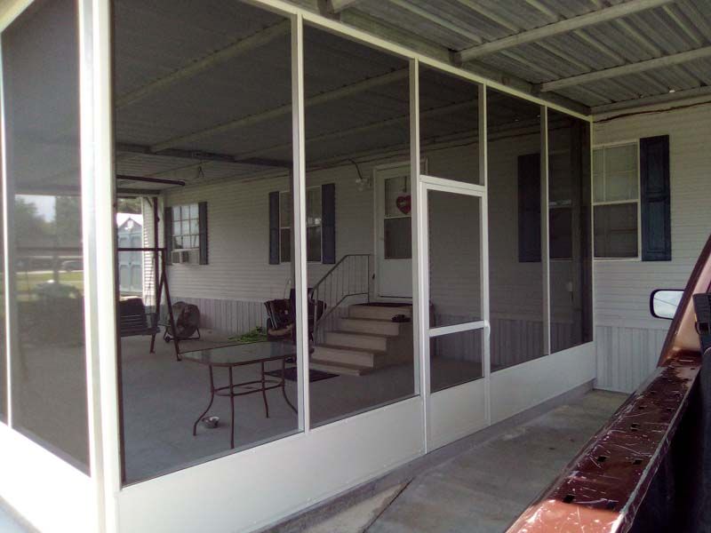Screened porch with white-framed windows, patio chairs, and a small table beside a concrete walkway.