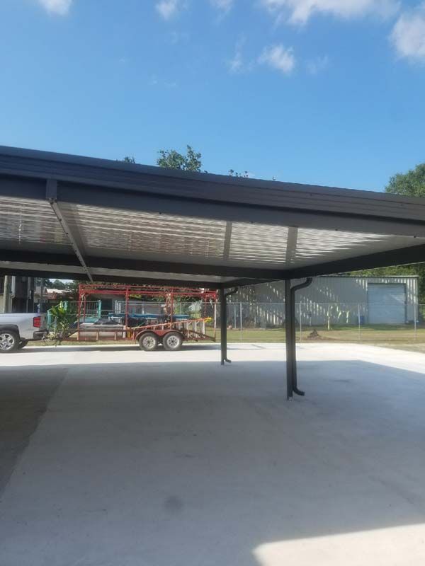 Open-air metal carport with concrete floor under a blue sky; utility trailer and truck in the background.