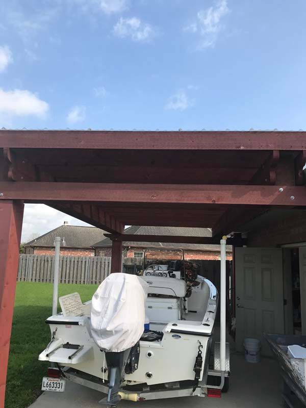 White boat on trailer parked under a red carport in a backyard with a fence and house nearby