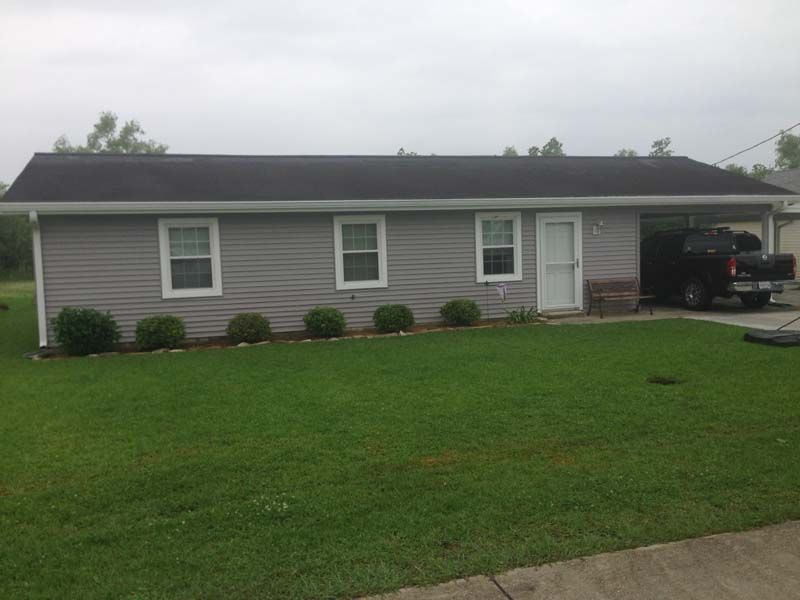 Single-story gray house with white trim, green lawn, and carport on the right under an overcast sky