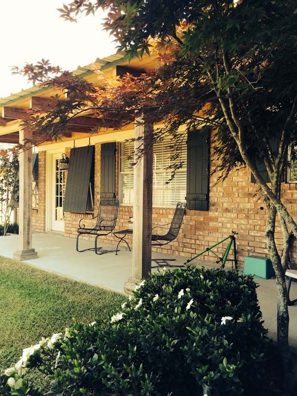 Front porch of a brick house with rocking chairs, white columns, and trees casting shade over the lawn.