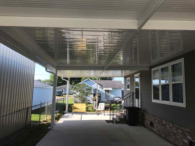Covered driveway beside a house, with a white fence and a sunlit yard in the background.