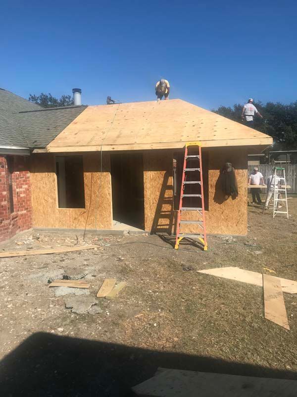 House under construction with plywood roof, ladder, and workers on a sunny day