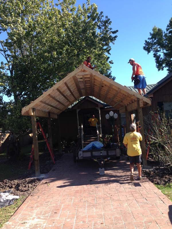 Two workers on a small roofed structure, one on the roof and one below, amid trees and a brick path.