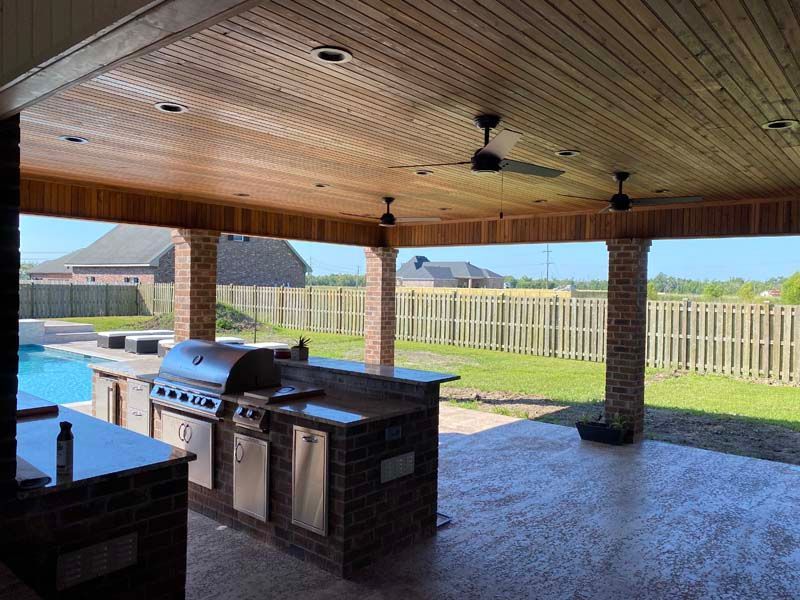 Covered patio with brick outdoor kitchen, ceiling fans, and backyard view through white fence.