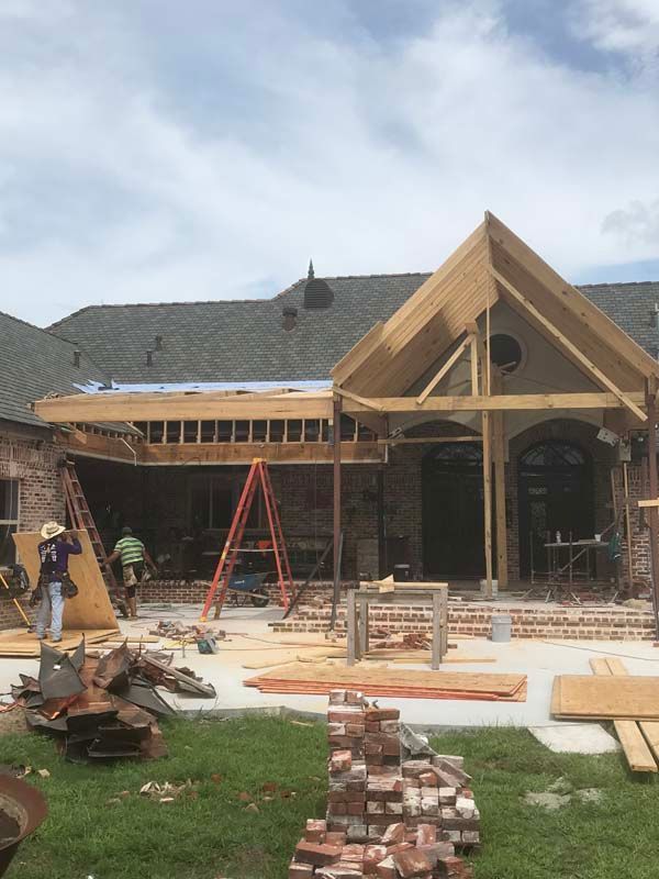 House under construction with wooden roof framing, ladders, brick stacks, and workers in a front yard