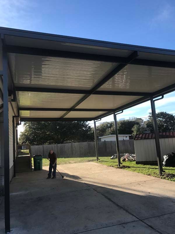 Covered driveway with a metal carport, concrete ground, fence, and a person standing in the shade.