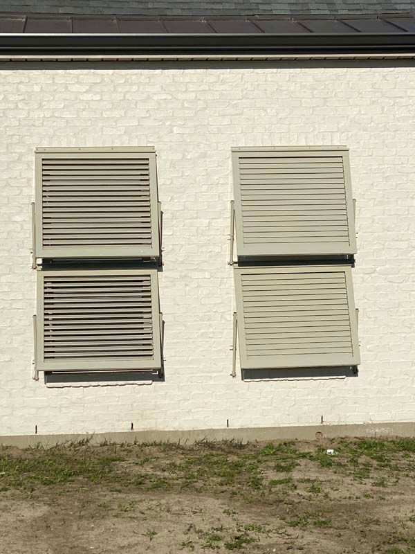 Four white louvered vents on a white brick wall above a dirt patch.