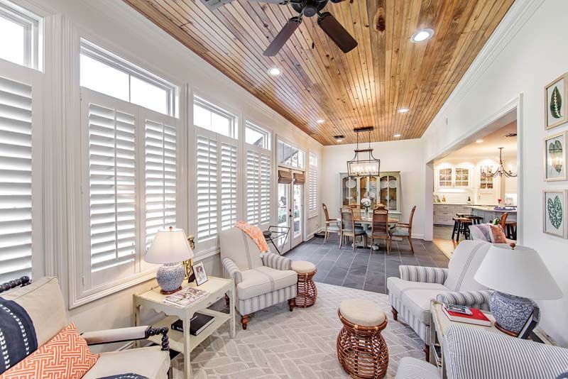 Bright sunroom with white shutters, striped armchairs, wood ceiling, and a small round table