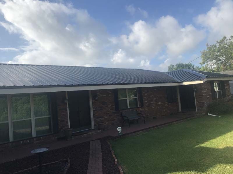 Single-story brick house with a metal roof, front porch, and green lawn under a cloudy sky