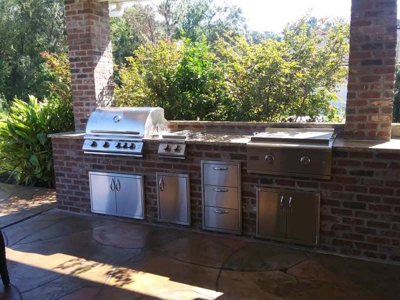 Outdoor brick patio kitchen with stainless steel grill, sink, and cabinet storage under a covered structure