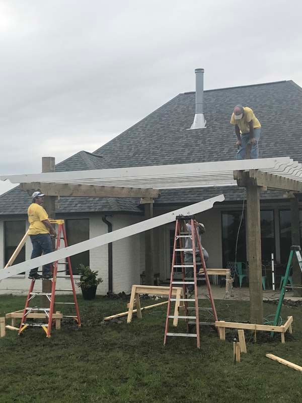 Workers installing a white patio cover on a house, with ladders and sawhorses in a backyard.