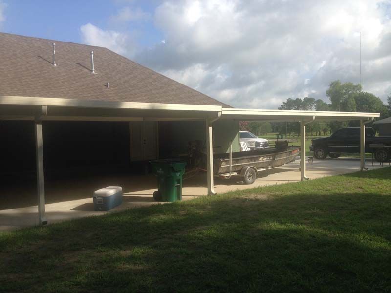 Covered carport beside a house with a parked truck, green trash bin, and grassy yard under cloudy sky