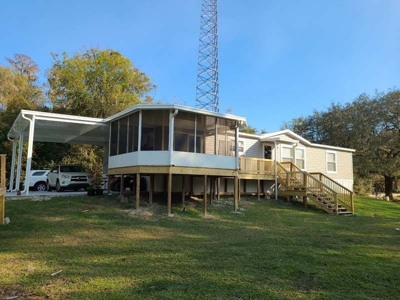 Rural mobile home with screened porch, carport, and wooden ramp on a grassy lot under blue sky