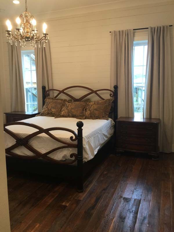 A cozy bedroom with a wooden bed, chandelier, dark hardwood floor, and windows with curtains.