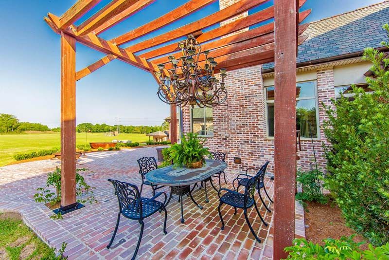 Brick patio with black metal table and chairs under a wooden pergola beside a house and green lawn