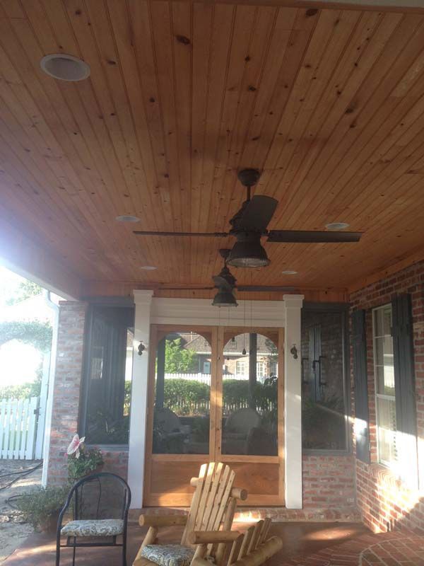 Covered porch with wood ceiling, ceiling fan, and chairs, facing a glass door and windows.