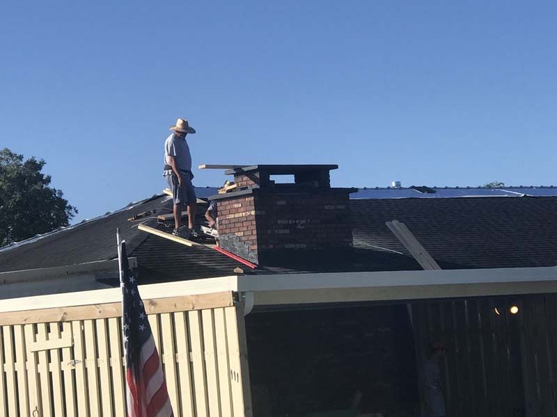 Person standing on a rooftop near a brick chimney, working on a house with an American flag in front.