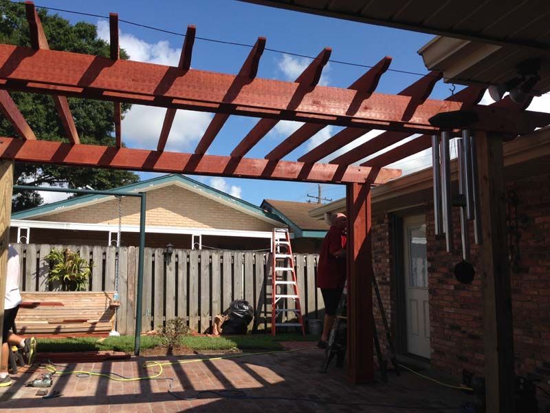 Backyard patio with red pergola beams, ladder, wooden fence, and brick house in sunlight