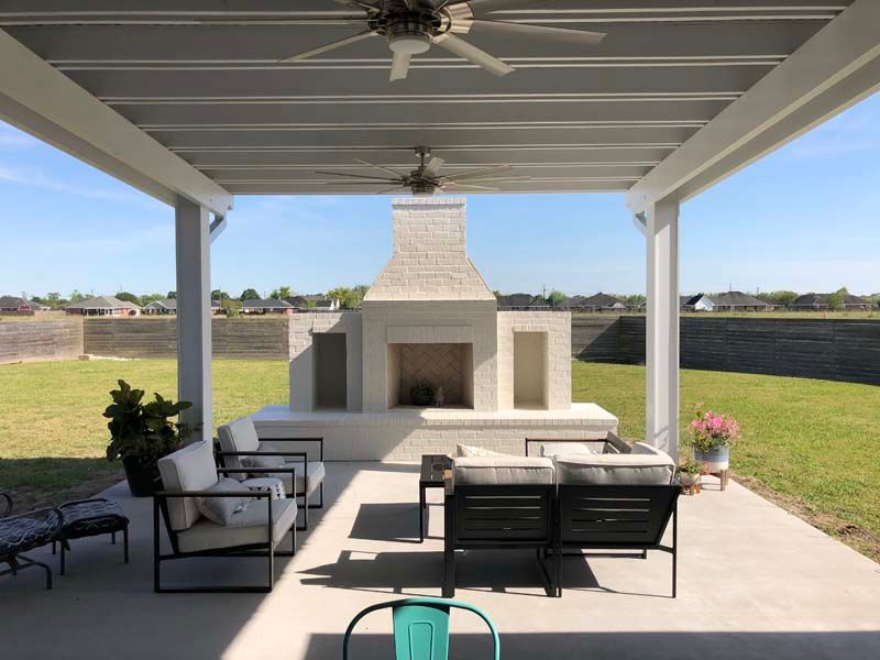 Covered patio with outdoor seating facing a large stone fireplace in a backyard.