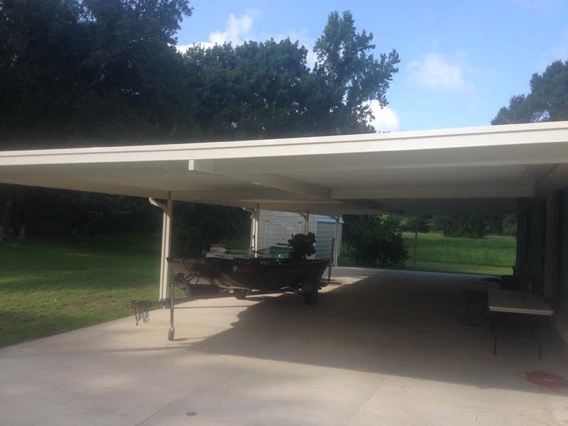 Carport with a parked trailer, concrete driveway, and trees in the background under a blue sky