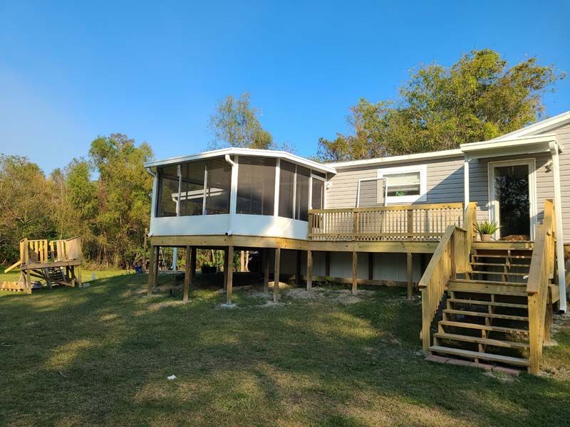 Raised beige house with screened porch, wooden deck, and stairs in a grassy yard under a blue sky