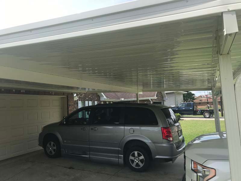 Silver SUV parked under a carport next to a beige garage, with a partial white car in the foreground.