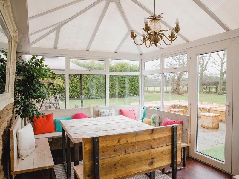 Sunlit conservatory dining area with wooden table, cushioned bench seating, and garden view