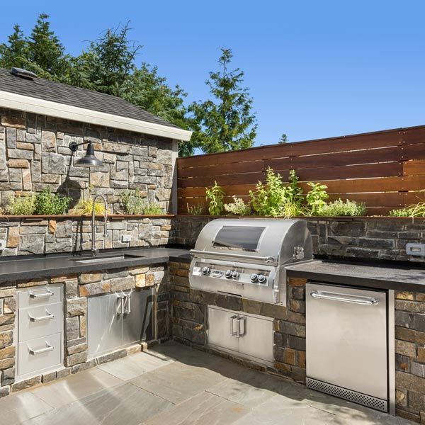 Stone outdoor kitchen with stainless grill, sink, cabinets, and fridge beside a stone wall and wooden fence