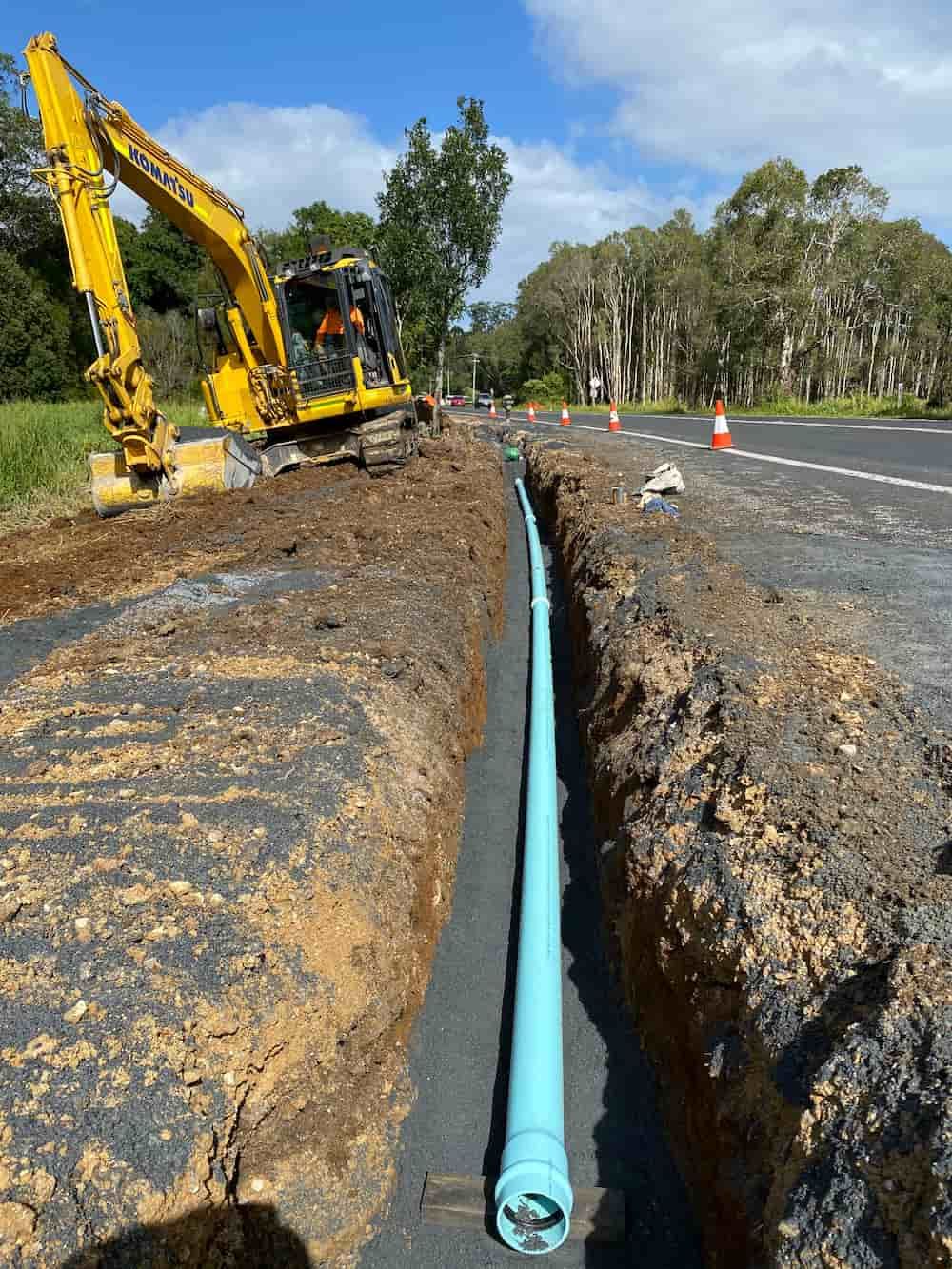 A Yellow Excavator Is Digging A Trench Next To A Green Pipe — J & M Bashforth & Sons In Brunswick Heads, NSW