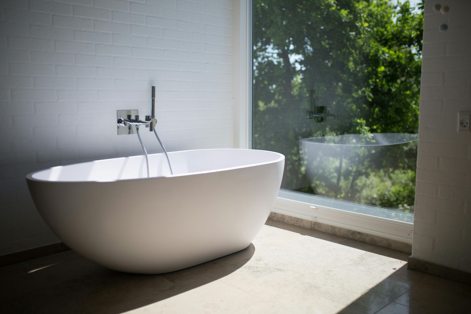 White oval bathtub near a large window overlooking greenery, in a bright bathroom with white walls and floor.
